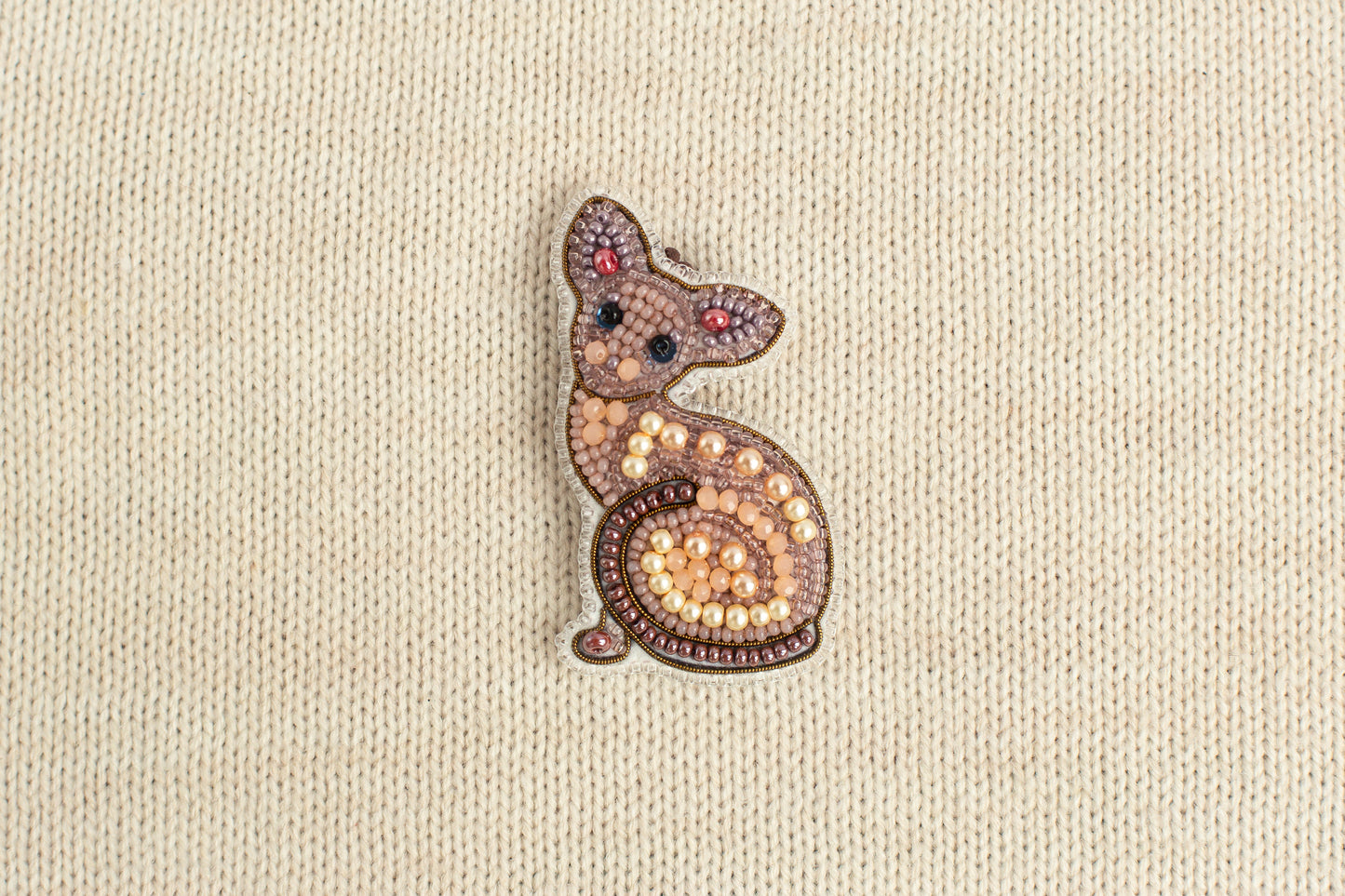 A decorative cat brooch made of beads and pearls is displayed against a beige fabric background.