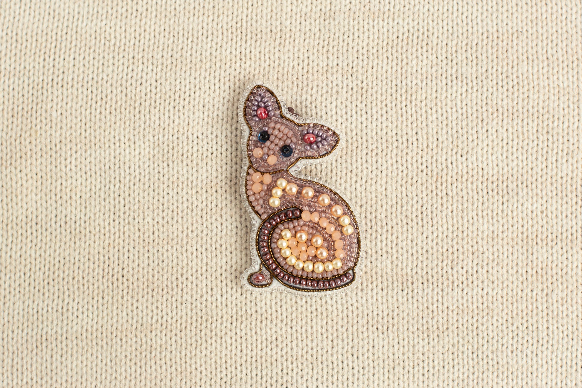 A decorative cat brooch made of beads and pearls is displayed against a beige fabric background.