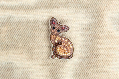 A decorative cat brooch made of beads and pearls is displayed against a beige fabric background.