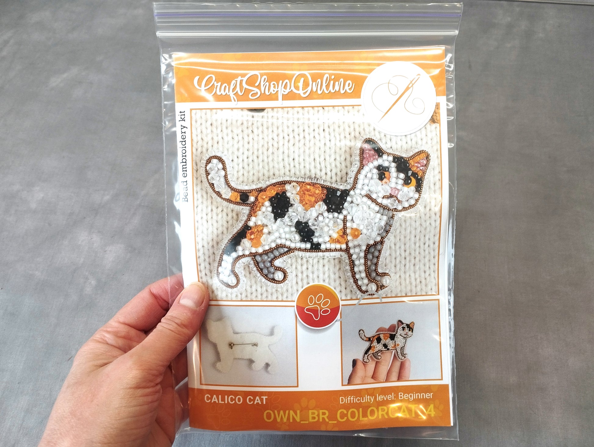 A hand is holding a package containing a kit for creating a calico cat embroidery.