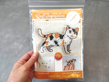 A hand is holding a package containing a kit for creating a calico cat embroidery.