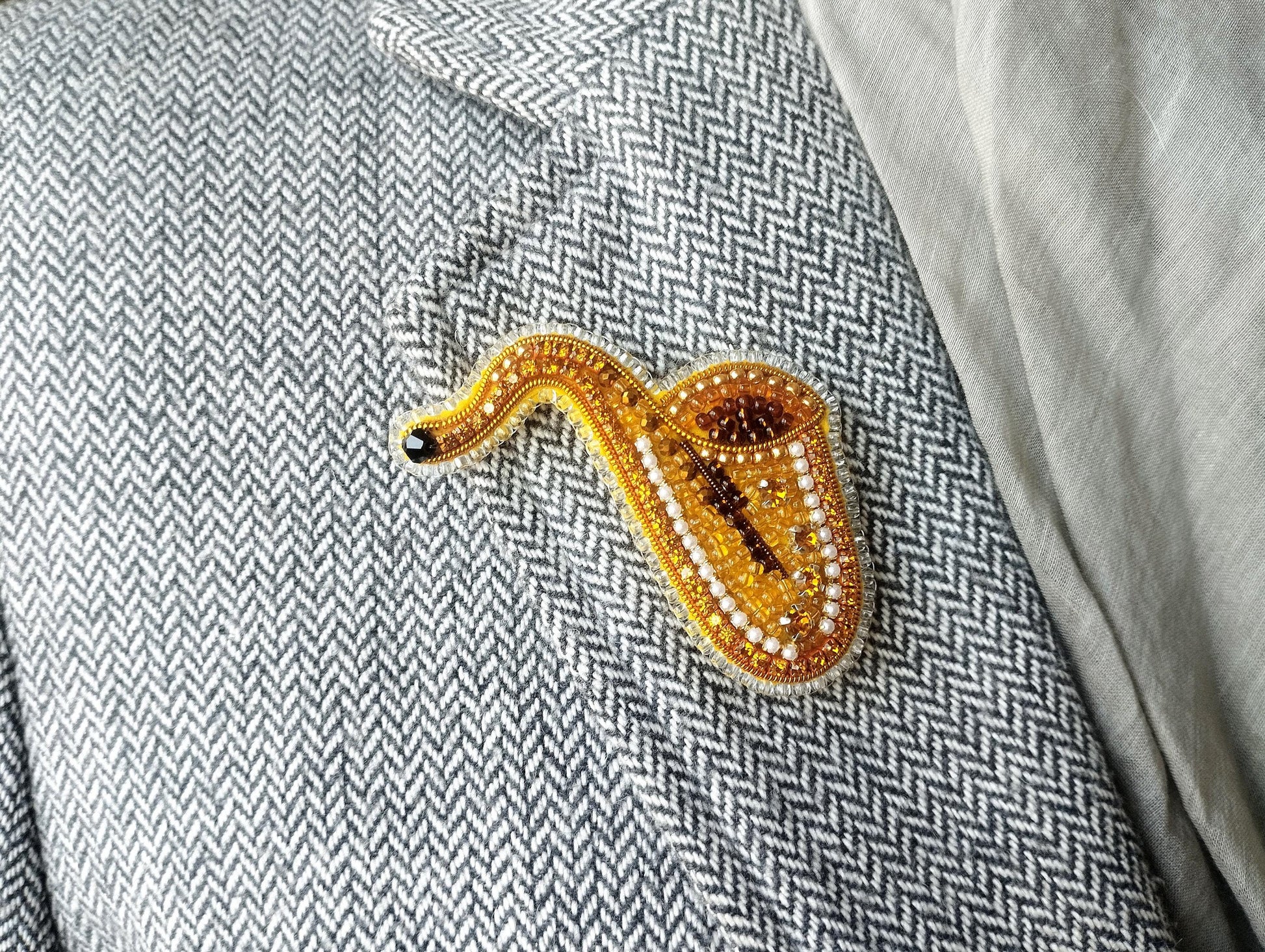 A close-up image of a gold and beaded brooch shaped like a saxophone, placed on a gray and white checkered fabric.