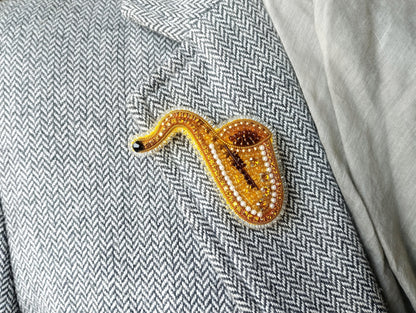 A close-up image of a gold and beaded brooch shaped like a saxophone, placed on a gray and white checkered fabric.