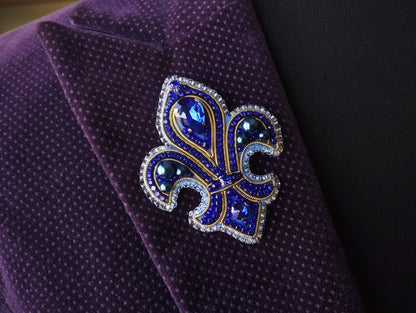 A close-up image of a purple velvet jacket with a decorative blue and gold brooch featuring a fleur-de-lis design and sparkling gemstones.