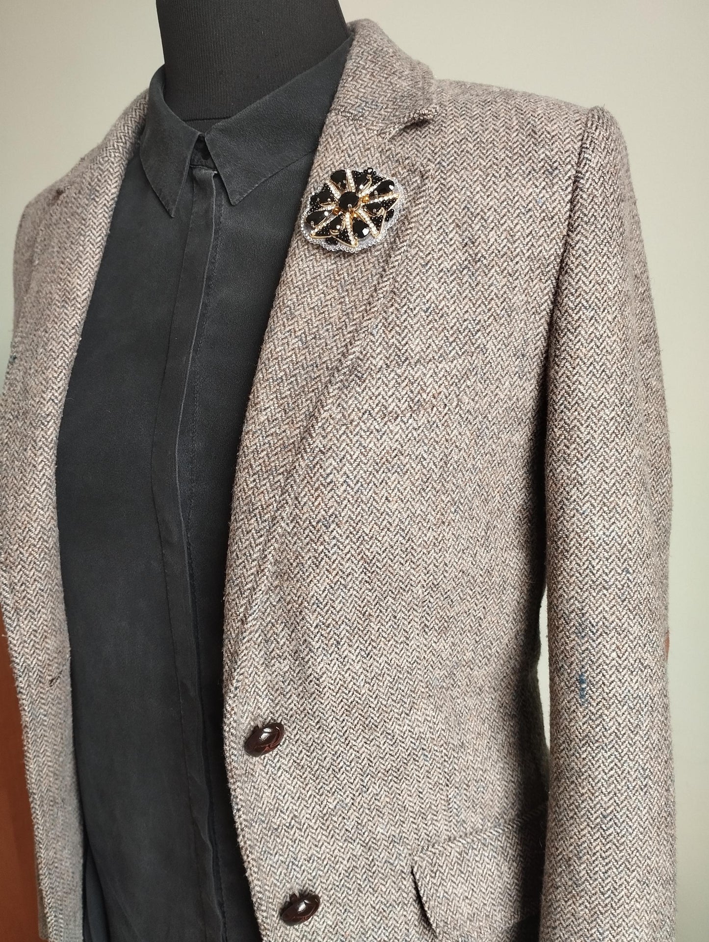 A mannequin wearing a gray tweed blazer with a black and gold brooch pinned to the lapel.