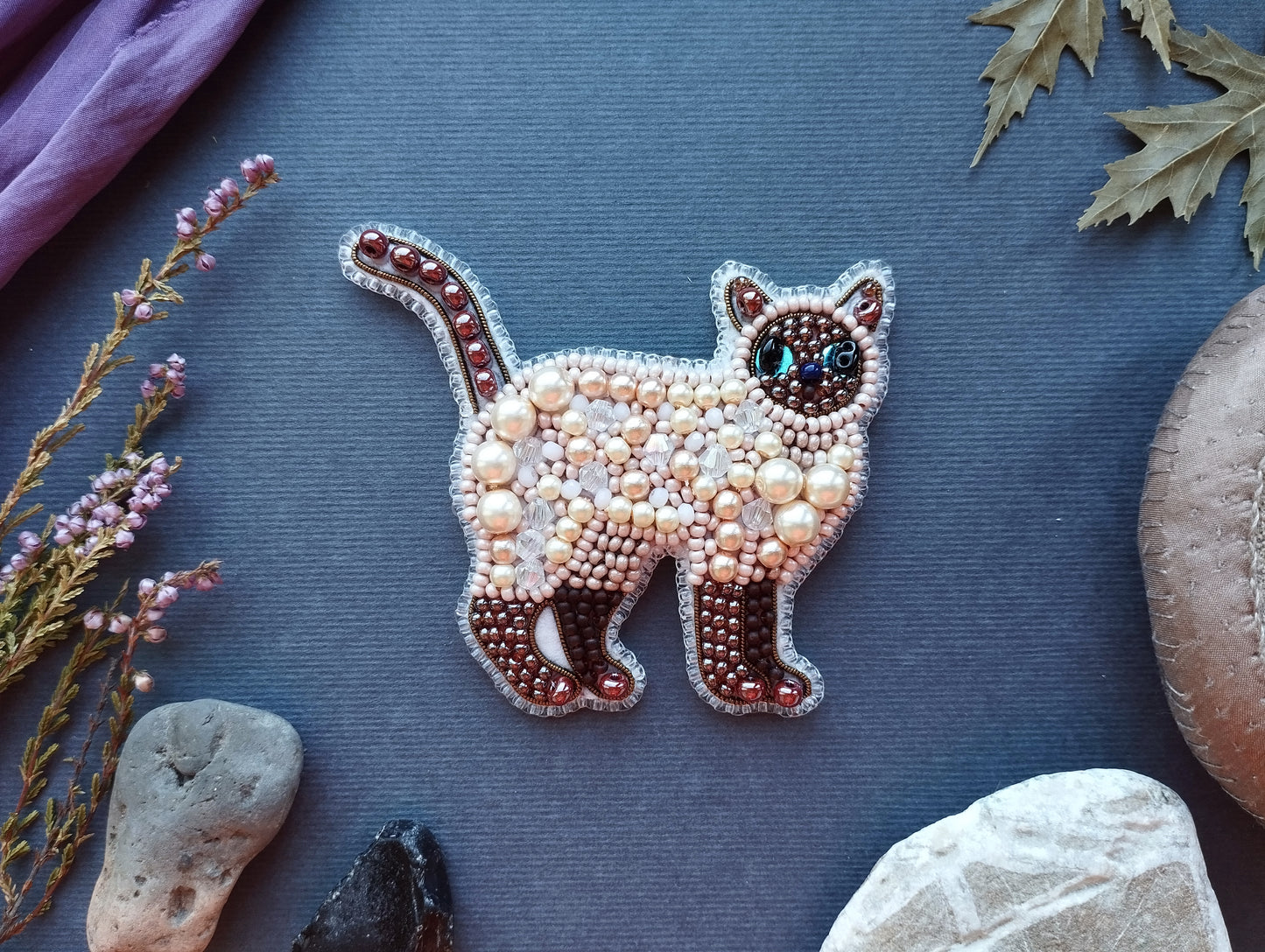 a decorative piece of embroidered cat-shaped jewelry, surrounded by various natural elements such as leaves, rocks, and flowers.