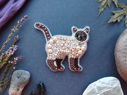 a decorative piece of embroidered cat-shaped jewelry, surrounded by various natural elements such as leaves, rocks, and flowers.