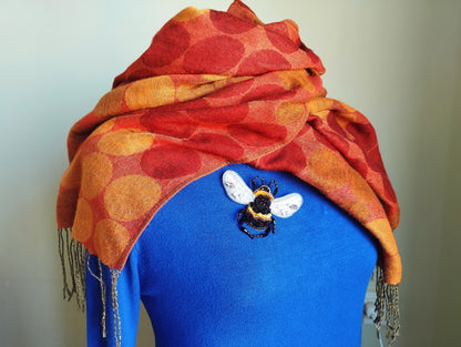 A woman wearing a blue sweater with a red and orange polka dot scarf draped over her shoulders, and a small embroidered bee pin on her chest.