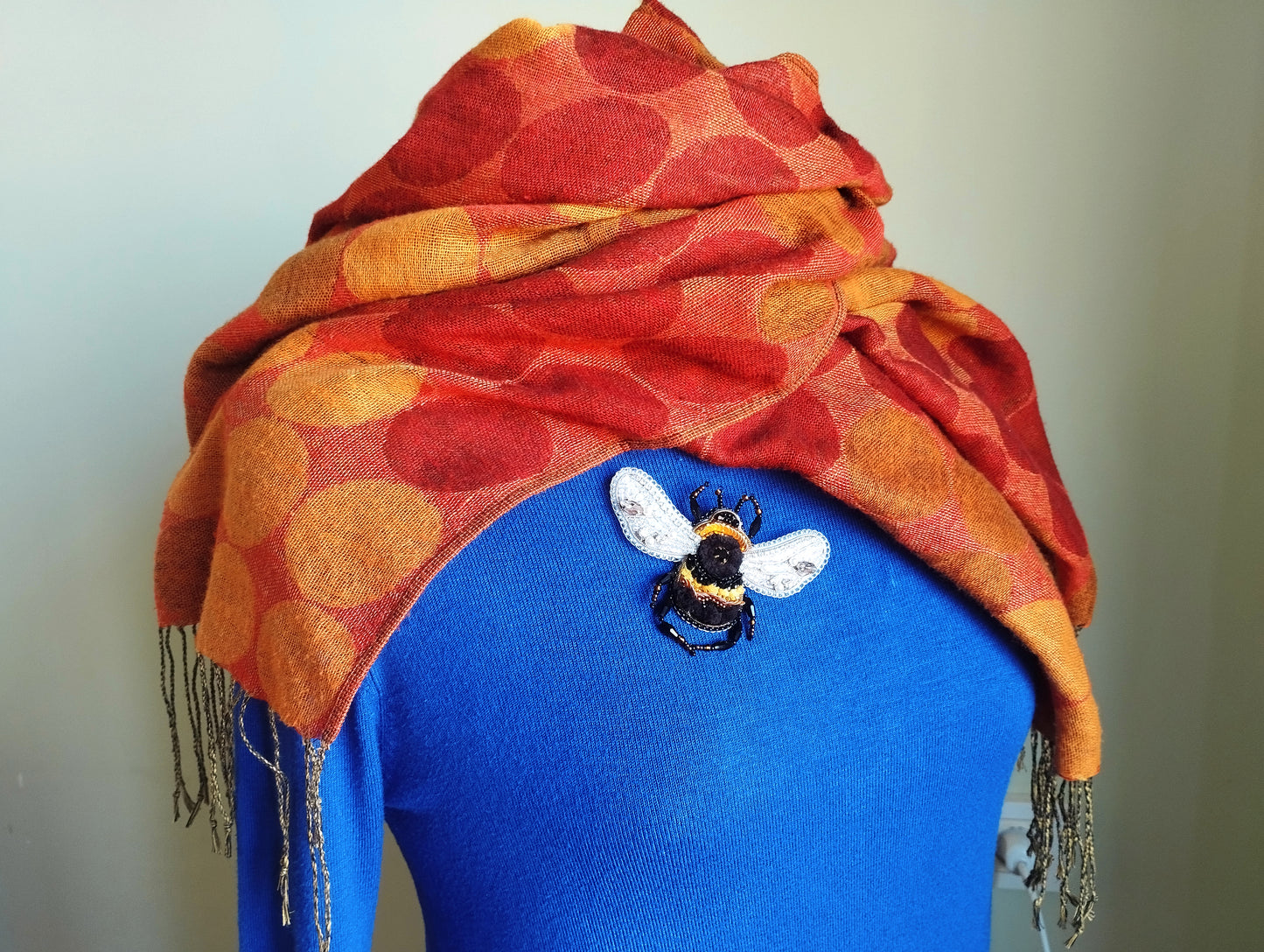 A woman wearing a blue sweater with a red and orange polka dot scarf draped over her shoulders, and a small embroidered bee pin on her chest.