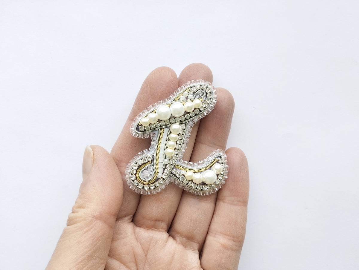 A hand holding a decorative letter 'L' made of beads and pearls.