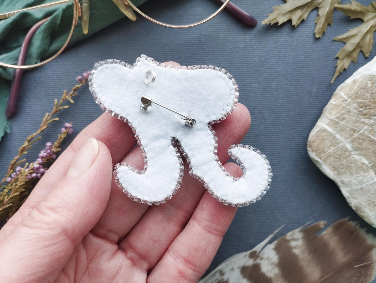 A hand holding a white felt elephant pin with a silver pin attached to it, surrounded by various natural elements such as leaves, feathers, and rocks.
