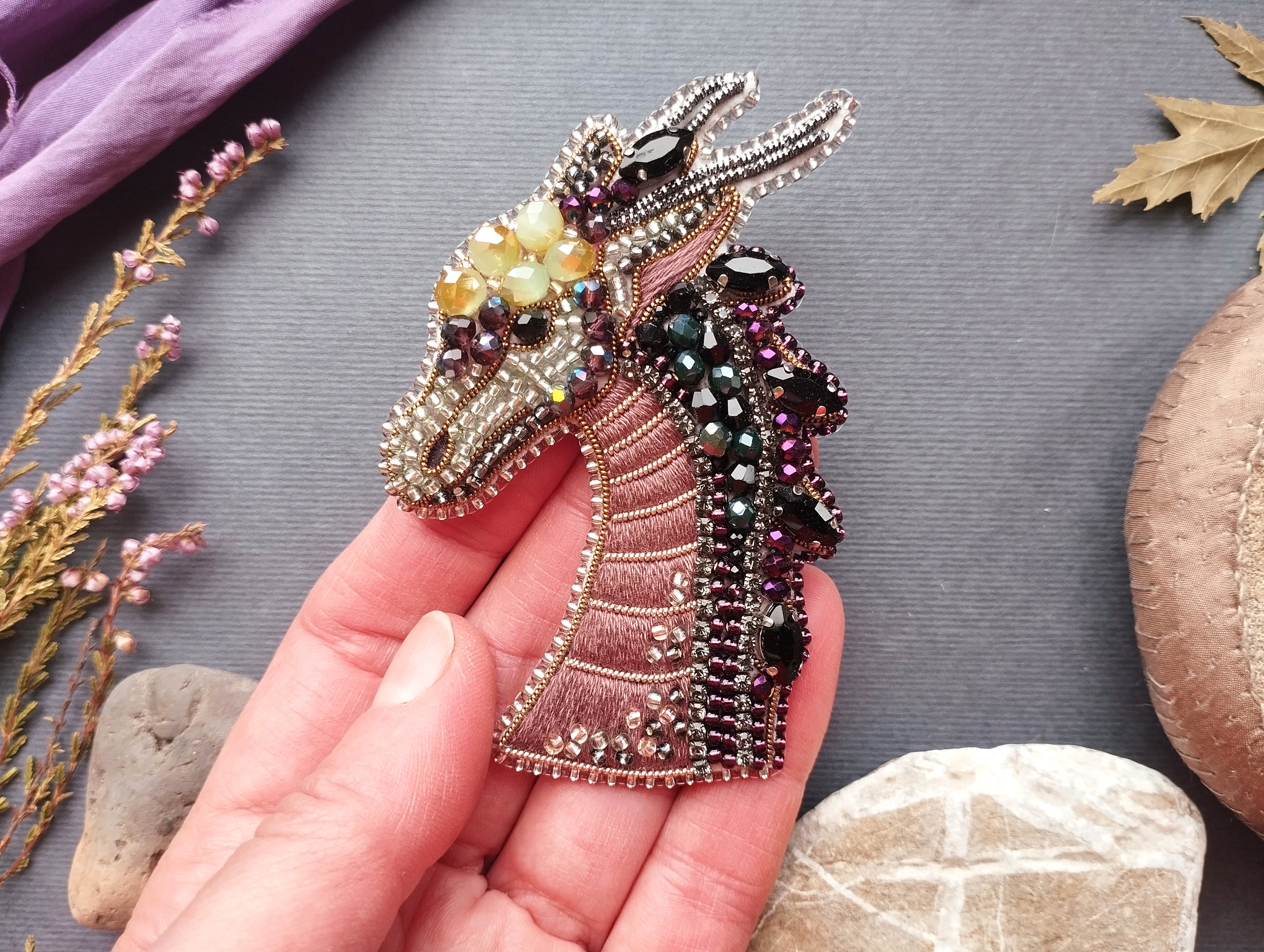 A hand holding a decorative beaded brooch in the shape of a dragon's head, with various colored beads and stones surrounding it.