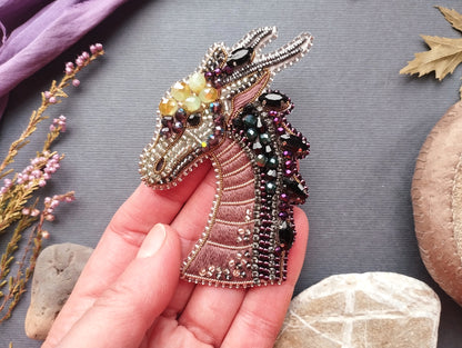 A hand holding a decorative beaded brooch in the shape of a dragon's head, with various colored beads and stones surrounding it.