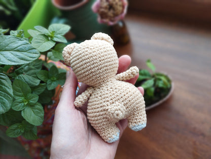 a hand holding a small crocheted teddy bear next to a potted plant