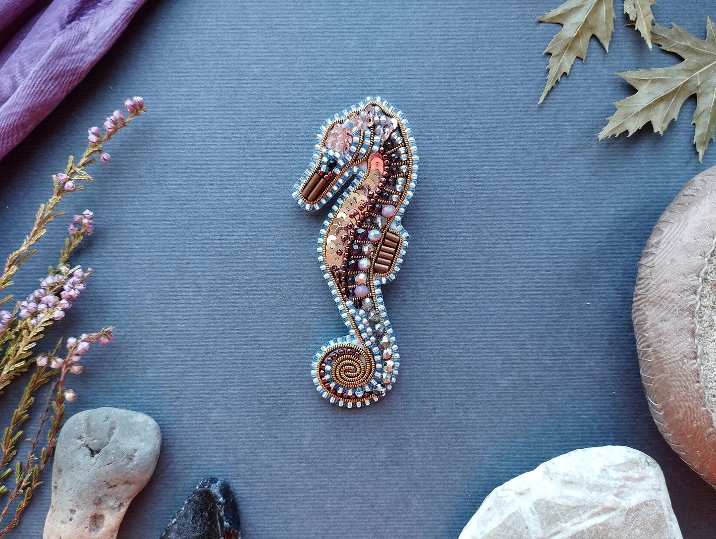 a decorative beaded seahorse brooch placed on a blue background, surrounded by various natural elements such as leaves, rocks, and flowers.