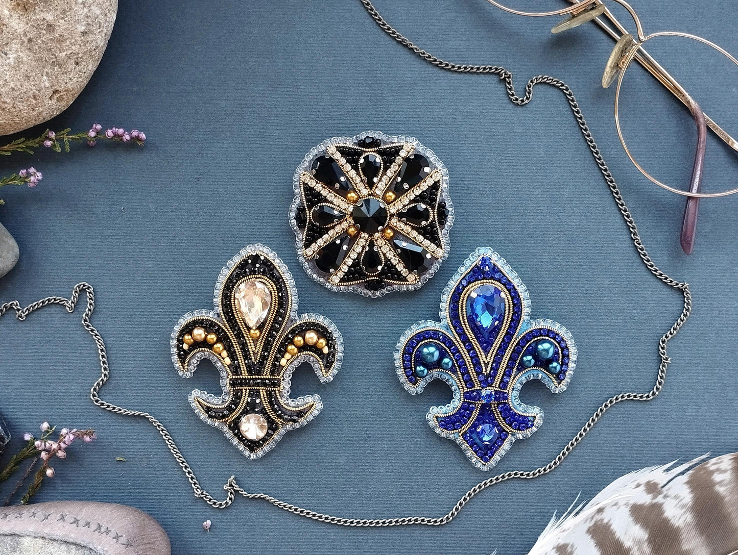 three ornate brooches with intricate designs and sparkling gemstones, arranged on a blue background.