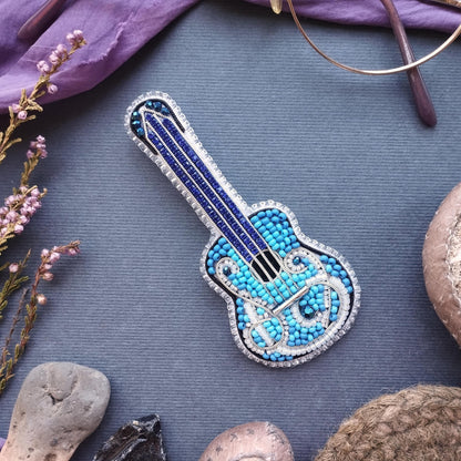 a blue and silver guitar brooch adorned with blue and silver beads, placed on a gray surface surrounded by various objects such as a pair of glasses, a flower, and a rock.