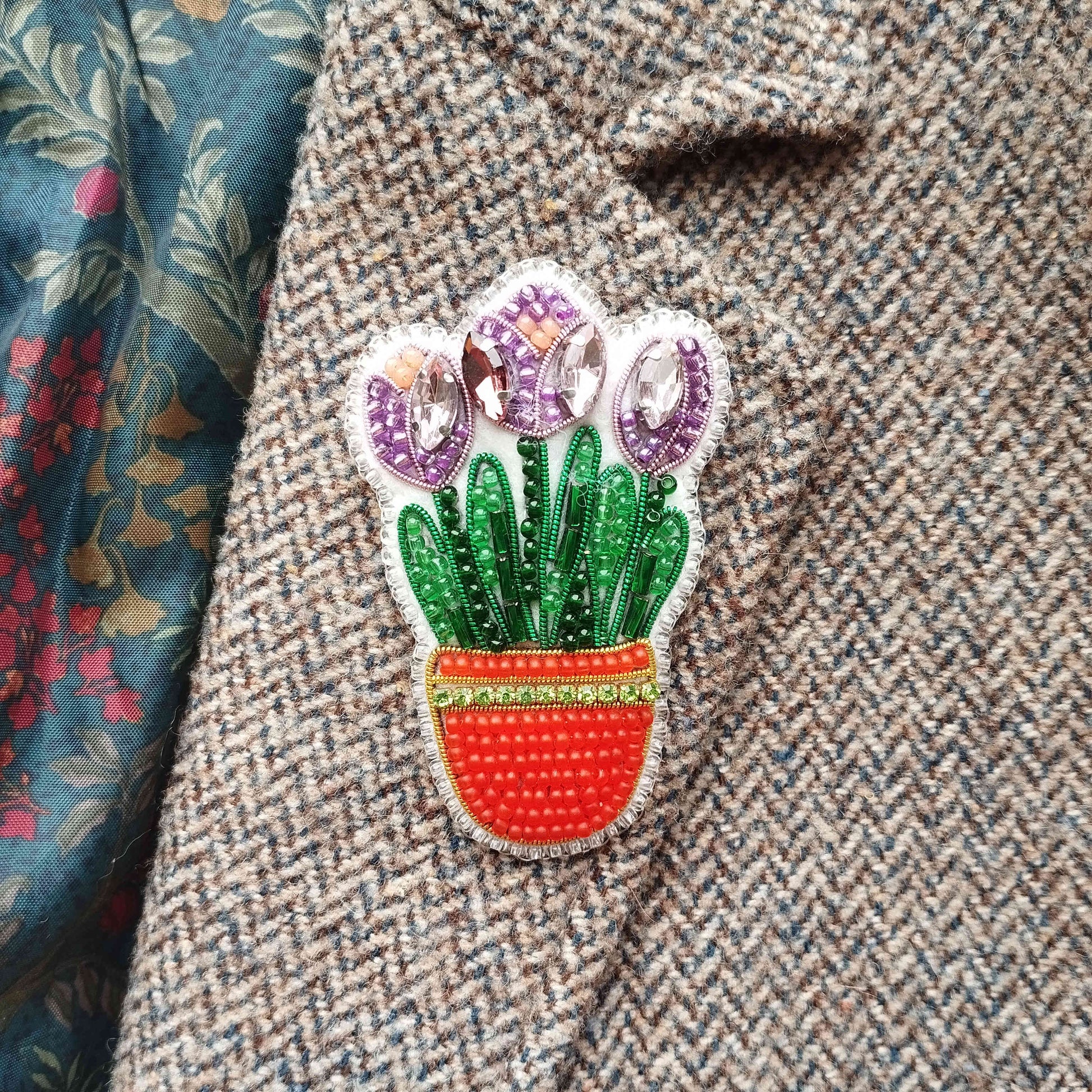 A close-up of a decorative pin or brooch featuring a potted plant with purple flowers, placed on a tweed jacket.