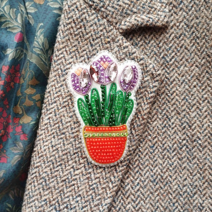 A close-up of a decorative pin or brooch featuring a potted plant with purple flowers, placed on a tweed jacket.