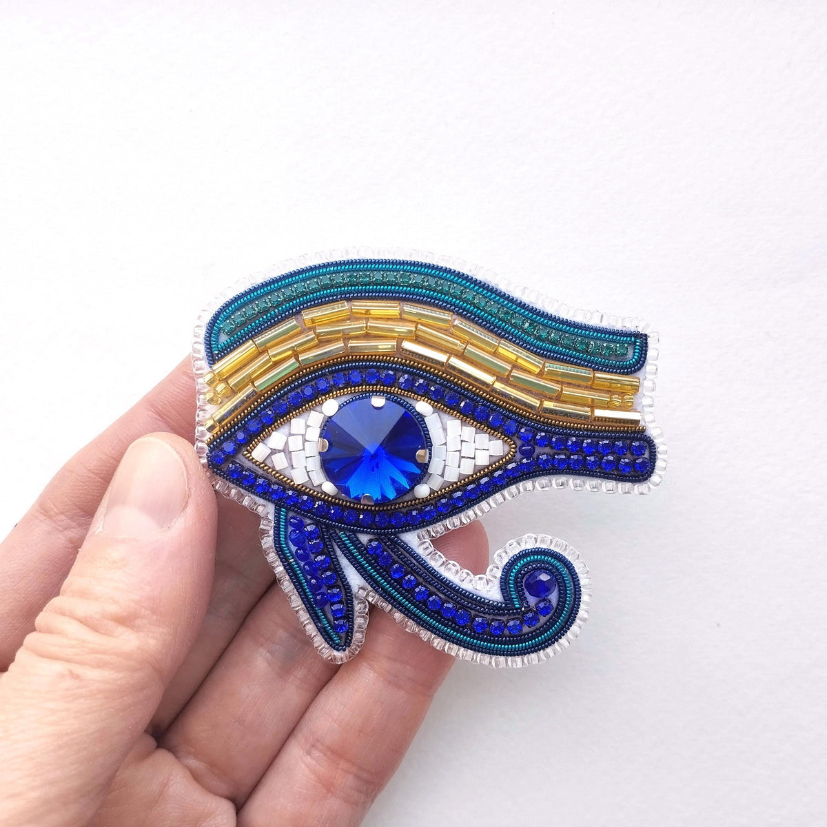 A hand holding a decorative piece featuring a large eye design with blue and gold elements, including a blue gemstone in the center.