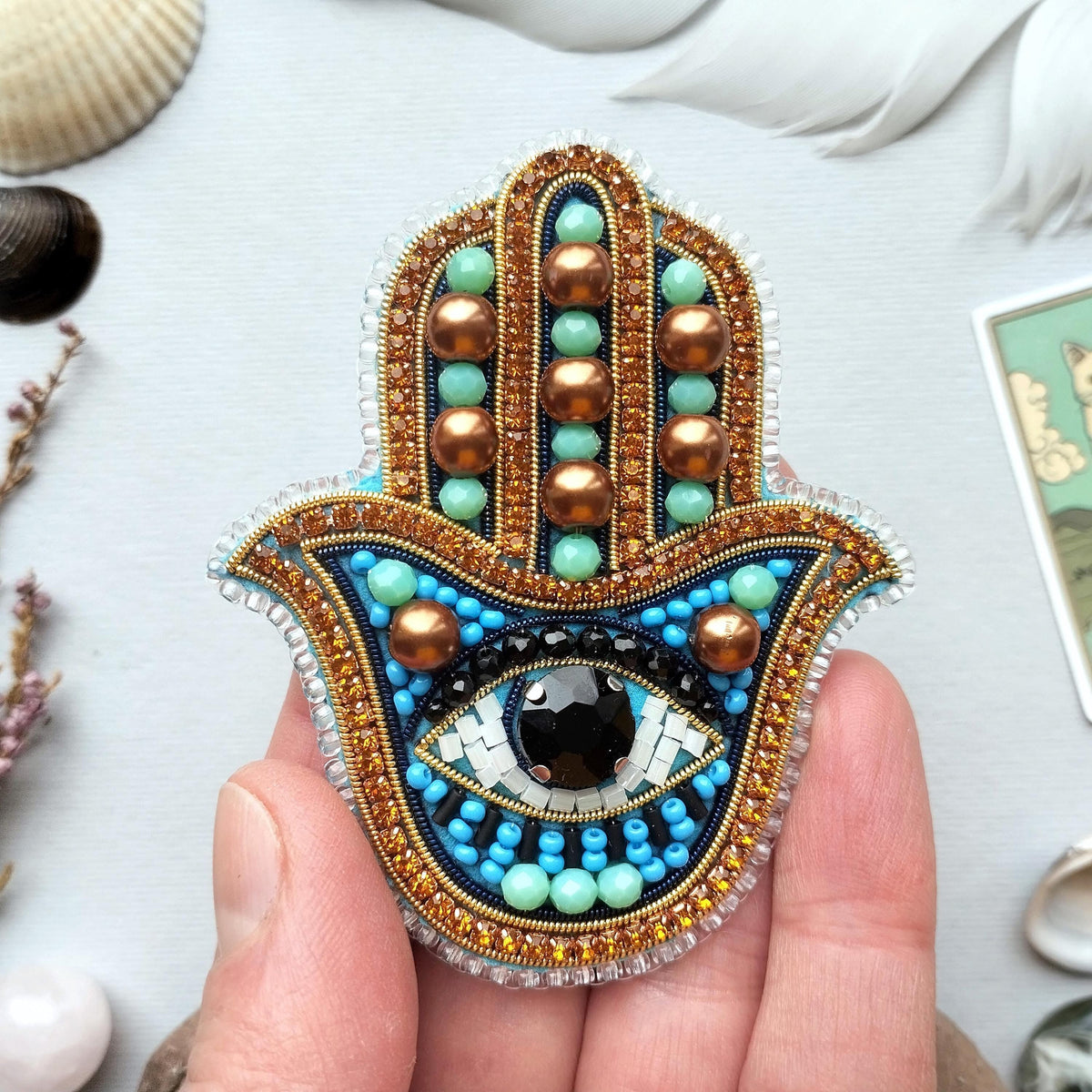 A hand holding a colorful, intricately beaded and embroidered Hamsa charm with a large eye design.