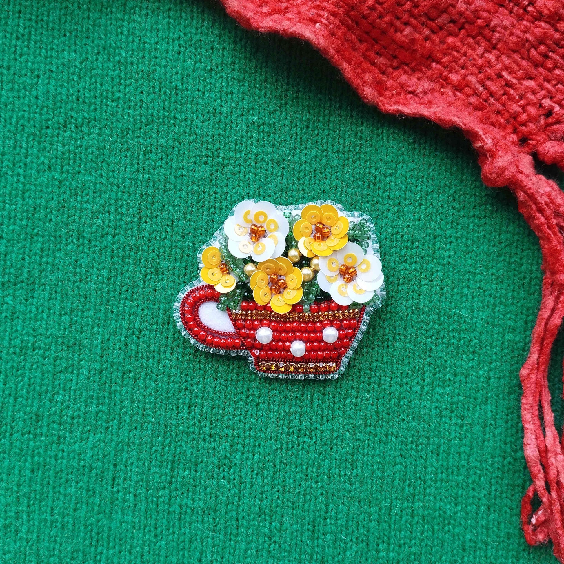 A red and white beaded brooch in the shape of a teacup, adorned with yellow and white flowers, is displayed against a green background.