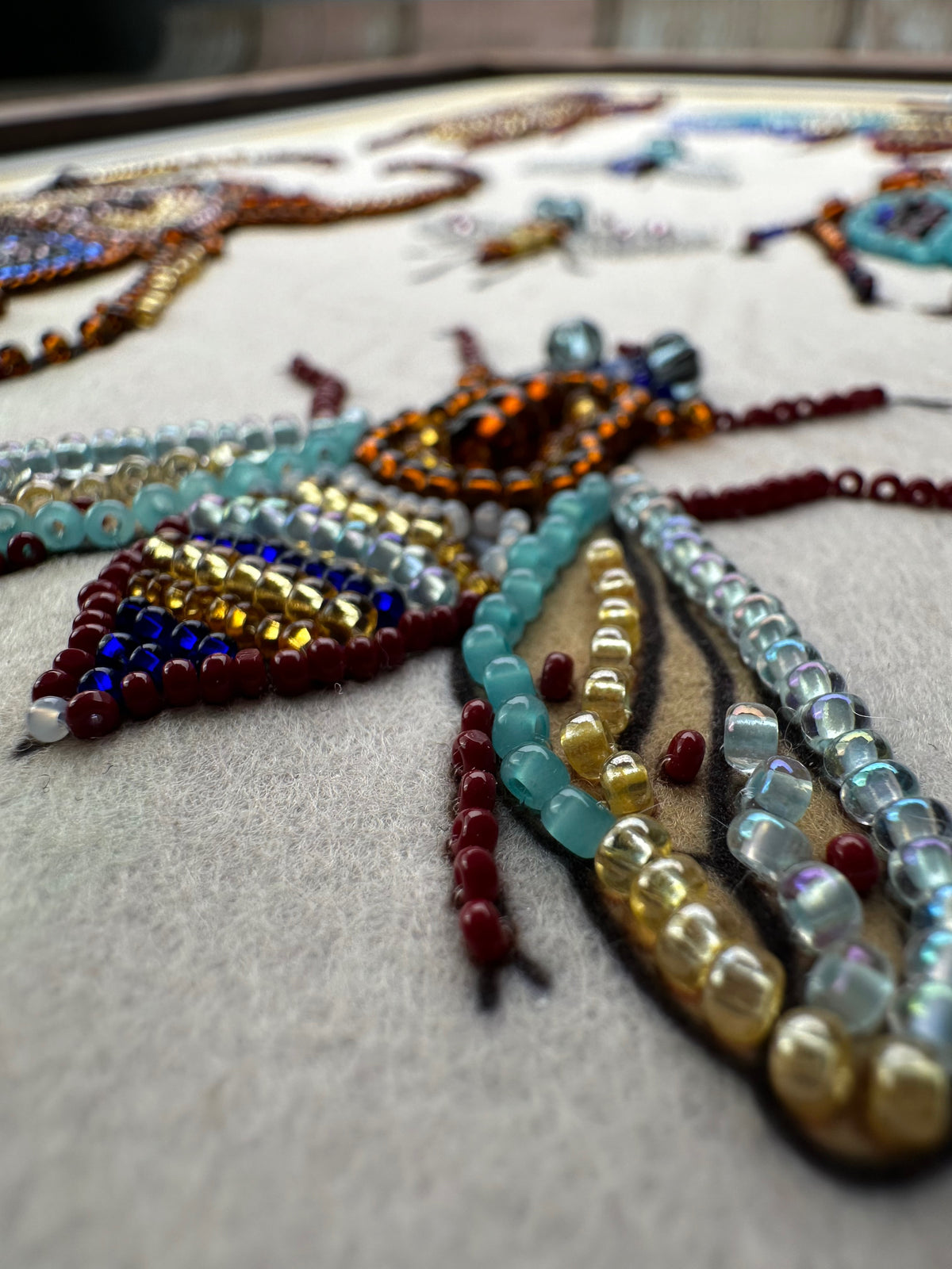 The image is a close-up of a detailed embroidered design featuring a colorful insect or bee-like creature made up of various beads, sequins, and other embellishments.