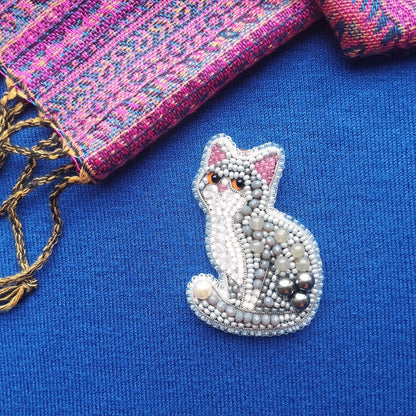 A beaded cat brooch is displayed on a blue fabric surface, with a pink and purple patterned fabric visible in the background.