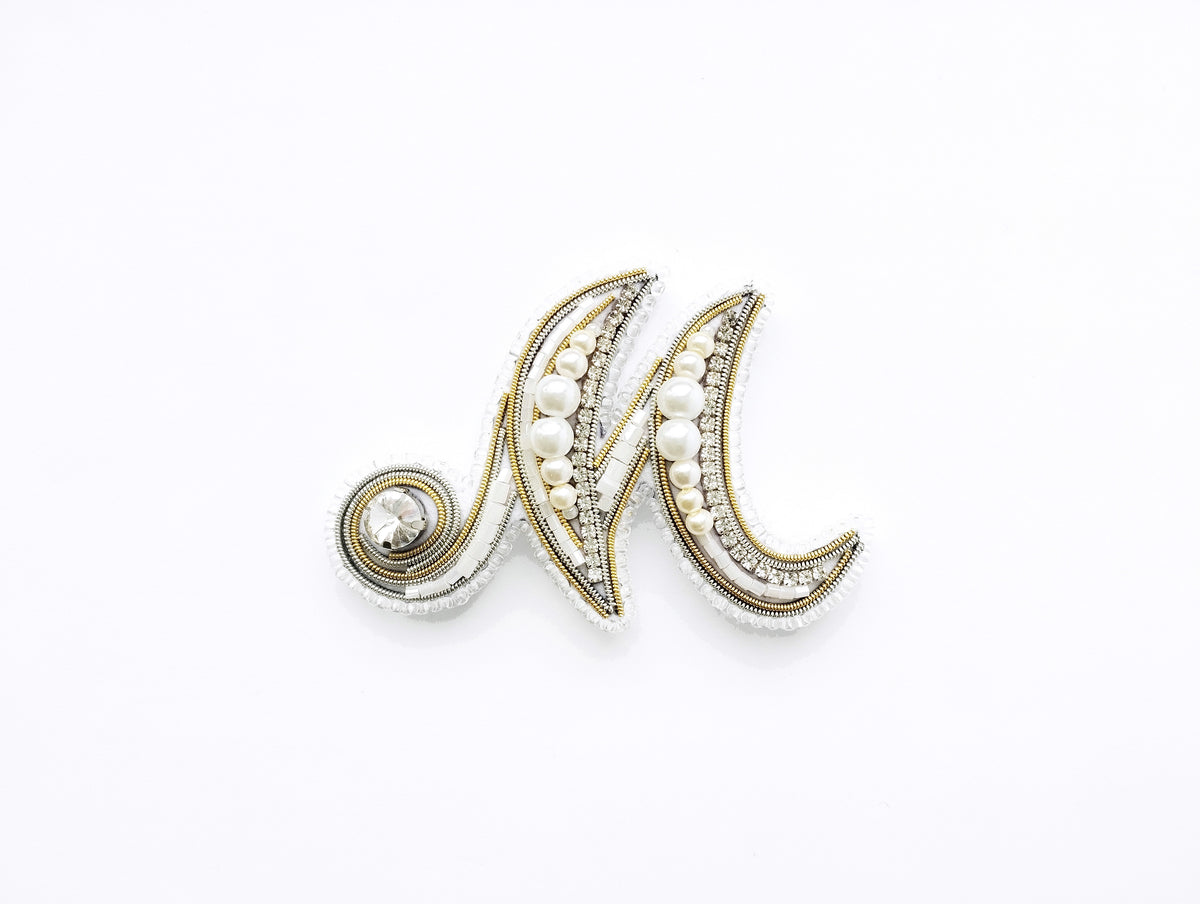 A decorative letter 'M' made of beads, pearls, and other embellishments, displayed against a white background.
