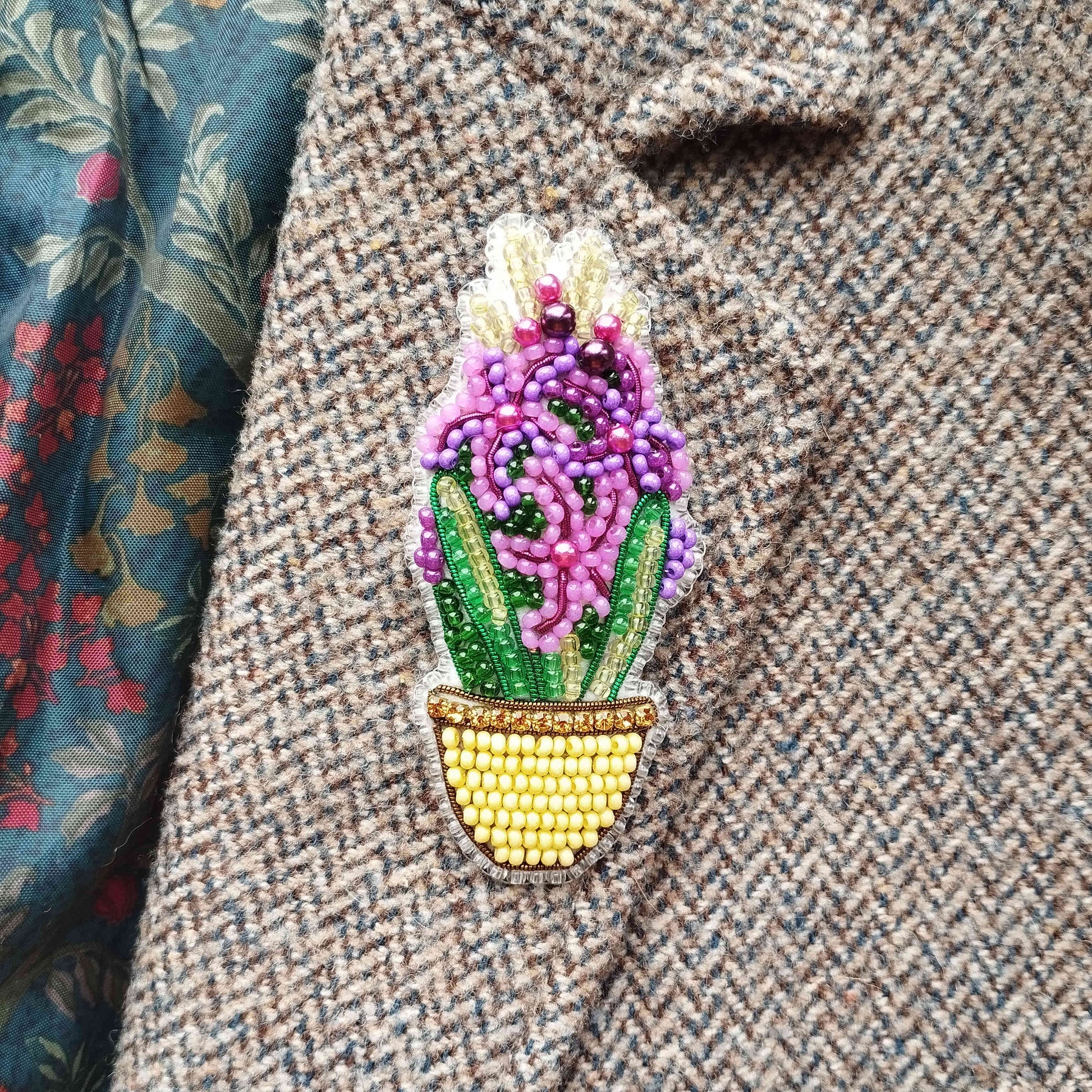 A close-up image of a decorative beaded pin or brooch featuring a floral design, placed on a tweed jacket or fabric with a floral pattern.