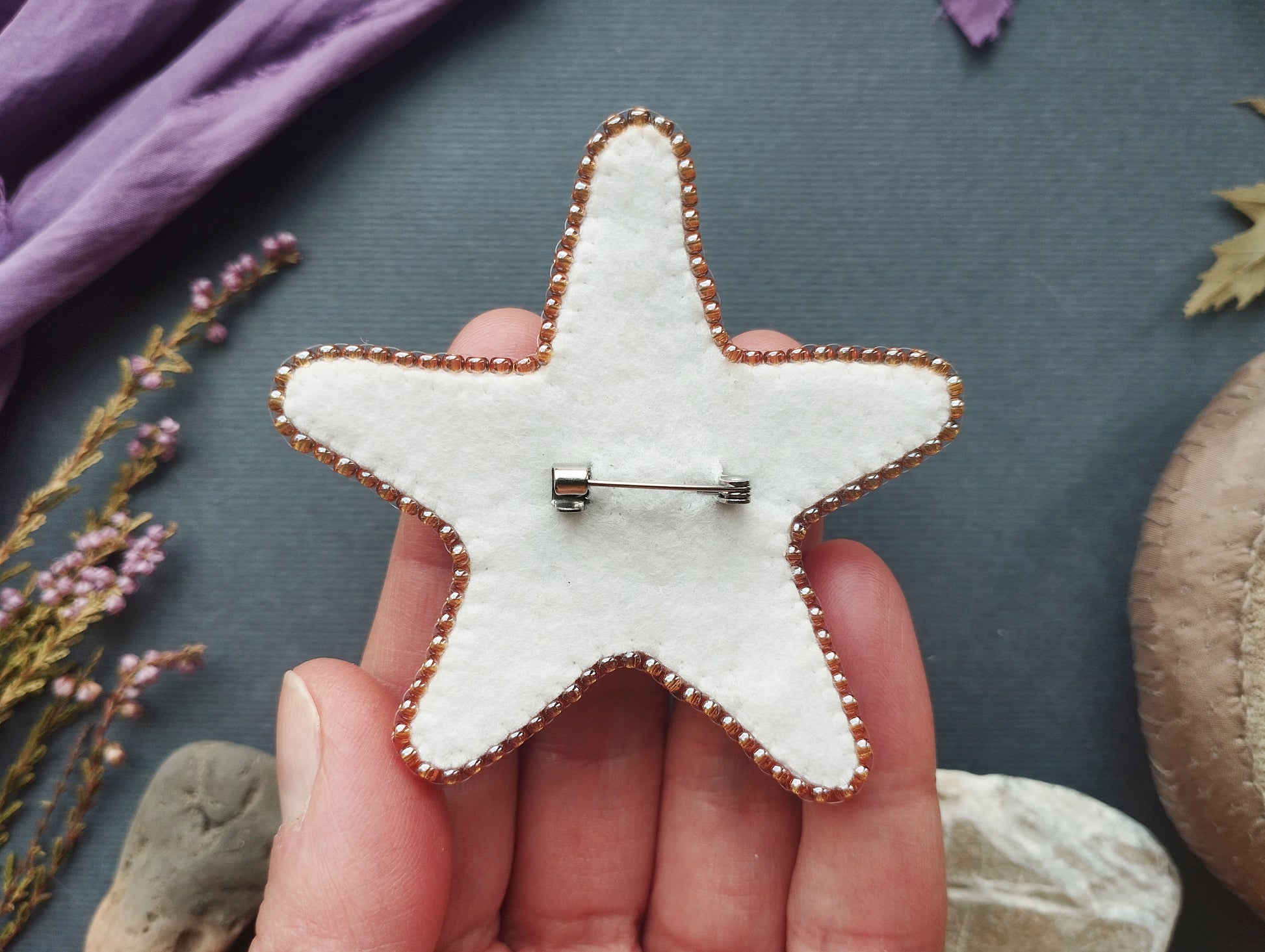 A hand holding a white star-shaped pin with a silver pinback on it, surrounded by various objects such as flowers, leaves, and rocks.