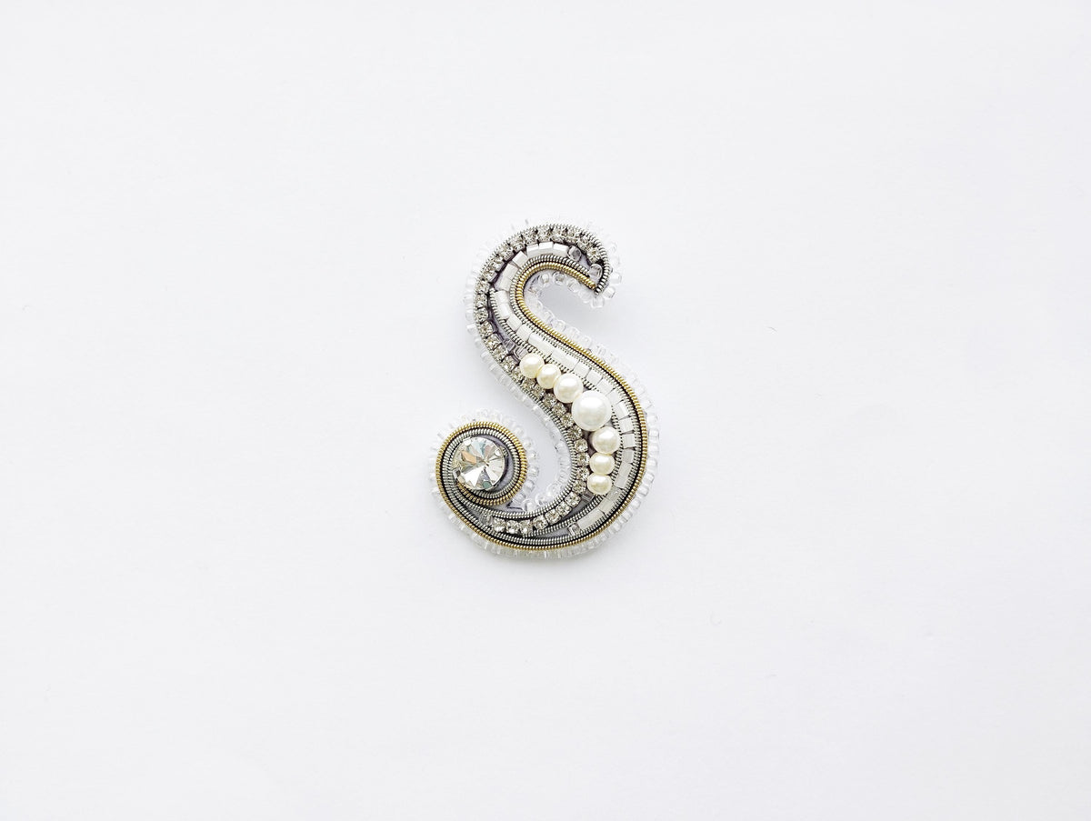 A decorative letter 'S' made of various materials, including pearls, beads, and crystals, against a plain white background.