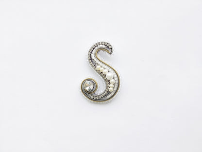 A decorative letter 'S' made of various materials, including pearls, beads, and crystals, against a plain white background.