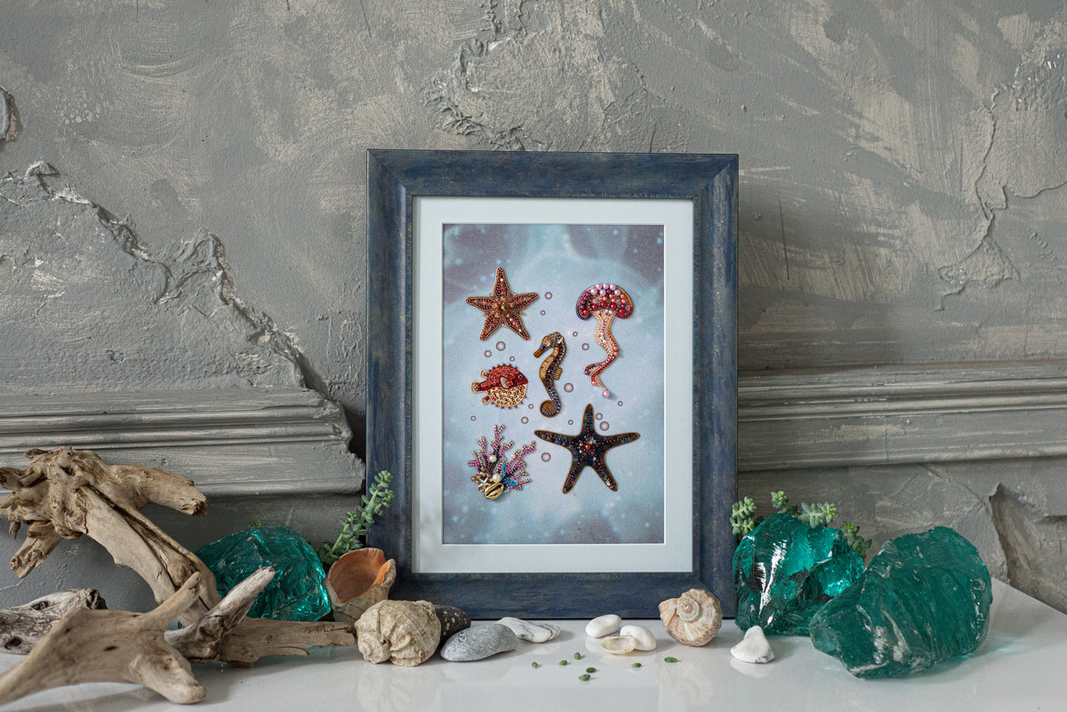 a framed artwork depicting various marine life, including starfish, a jellyfish, and a seahorse, set against a blue background. The artwork is surrounded by various natural elements, such as driftwood, rocks, and green glass, creating a serene and coastal atmosphere.