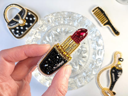 A hand holding a decorative piece featuring a red gemstone and black and gold embellishments, with other decorative pieces in the background.