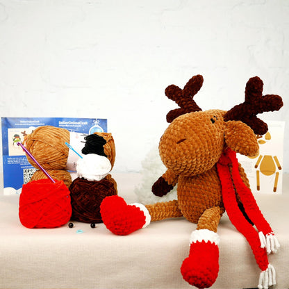 A brown plush reindeer toy sits on a white surface, wearing red knitted mittens and a red scarf. Next to the reindeer is a ball of yarn and a crochet hook, suggesting the toy is being crafted. In the background, there is a blue sign with white text, and a white card with a black and white illustration.