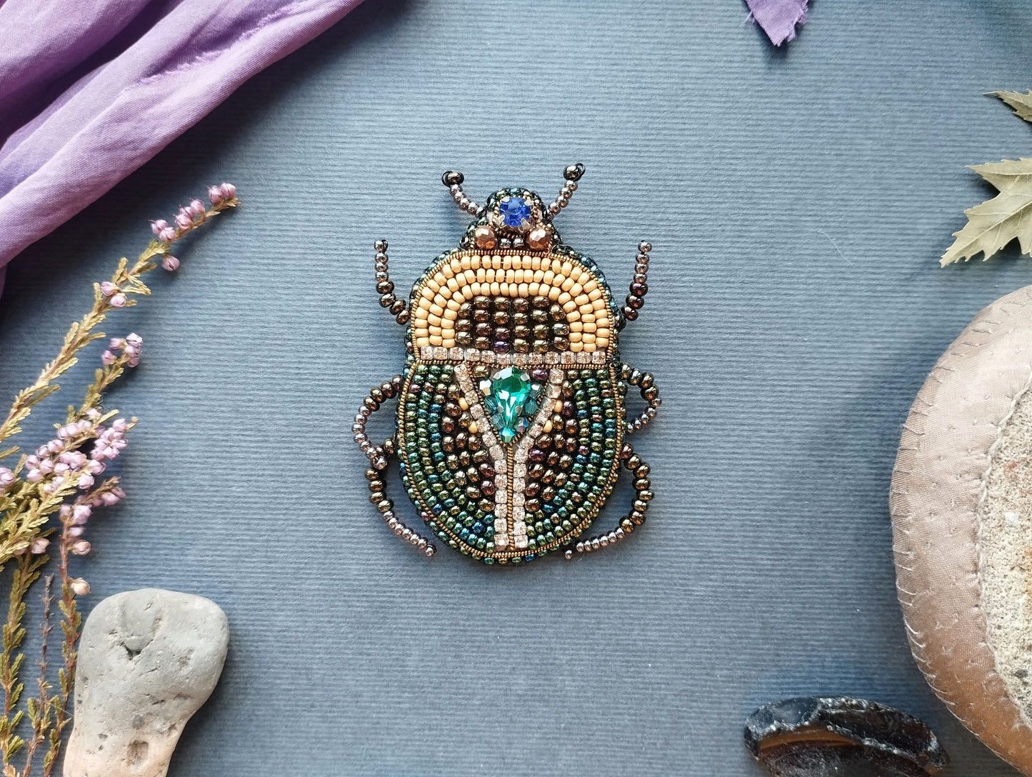 a detailed beaded insect brooch with a vibrant design, placed on a blue background. Surrounding the brooch are various natural elements, including a purple fabric, a small flower, and a rock.