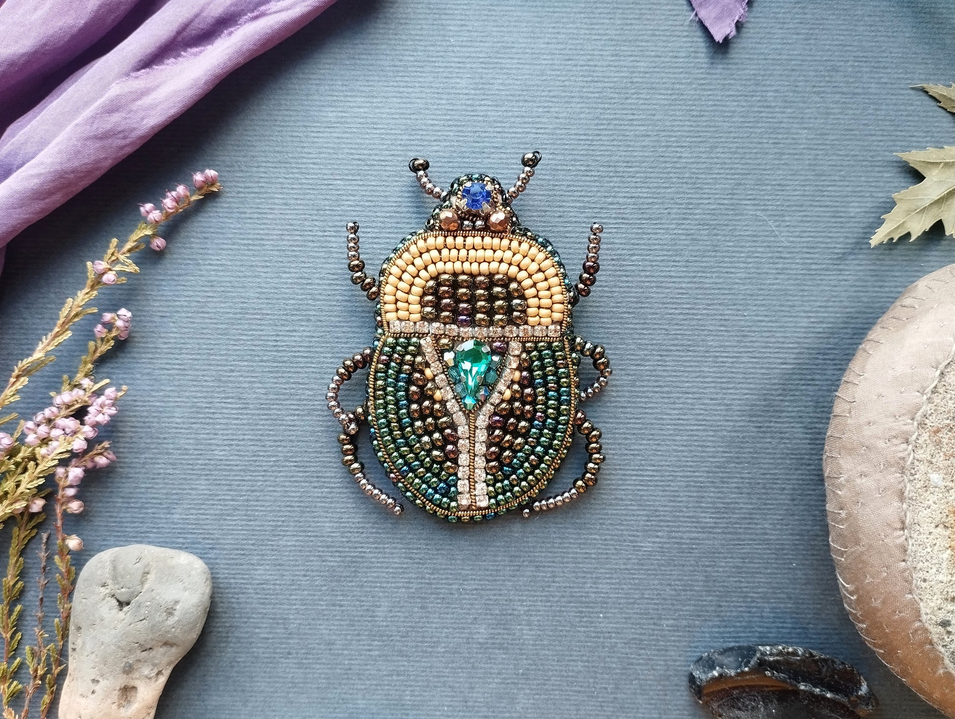 a detailed beaded insect brooch with a vibrant design, placed on a blue background. Surrounding the brooch are various natural elements, including a purple fabric, a small flower, and a rock.