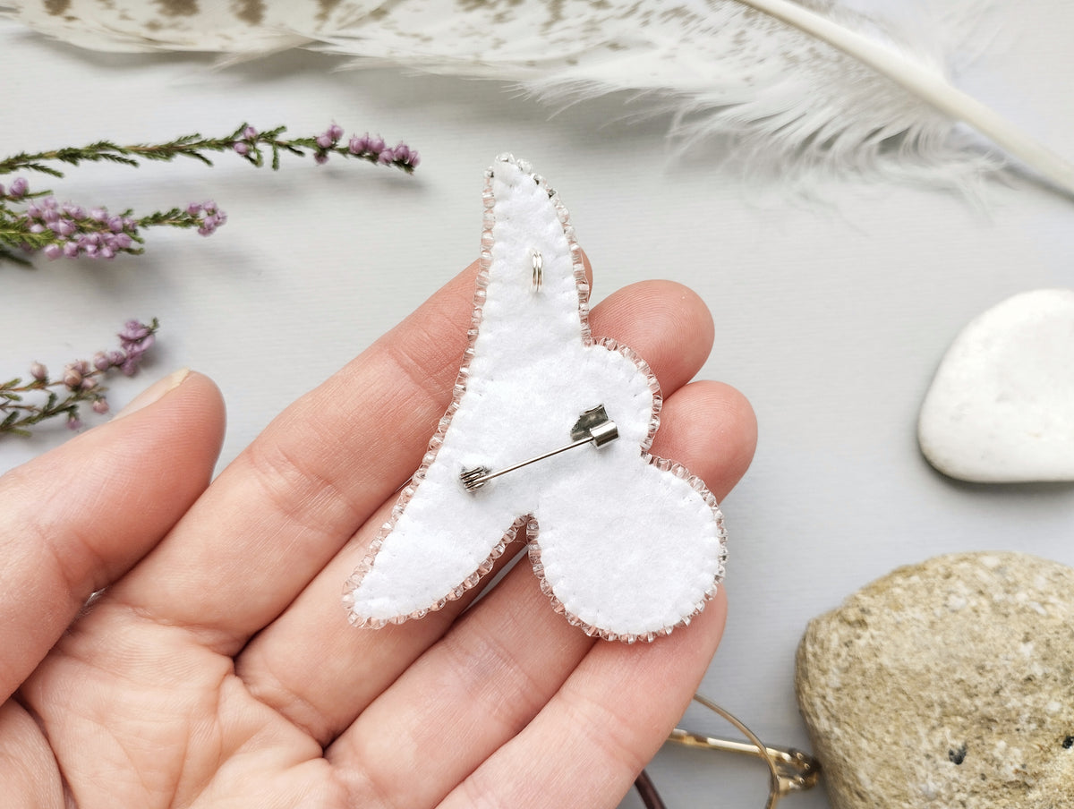 A hand holding a white felt pin or badge with a silver pin or clasp attached to it, surrounded by various natural elements such as feathers, flowers, and rocks.