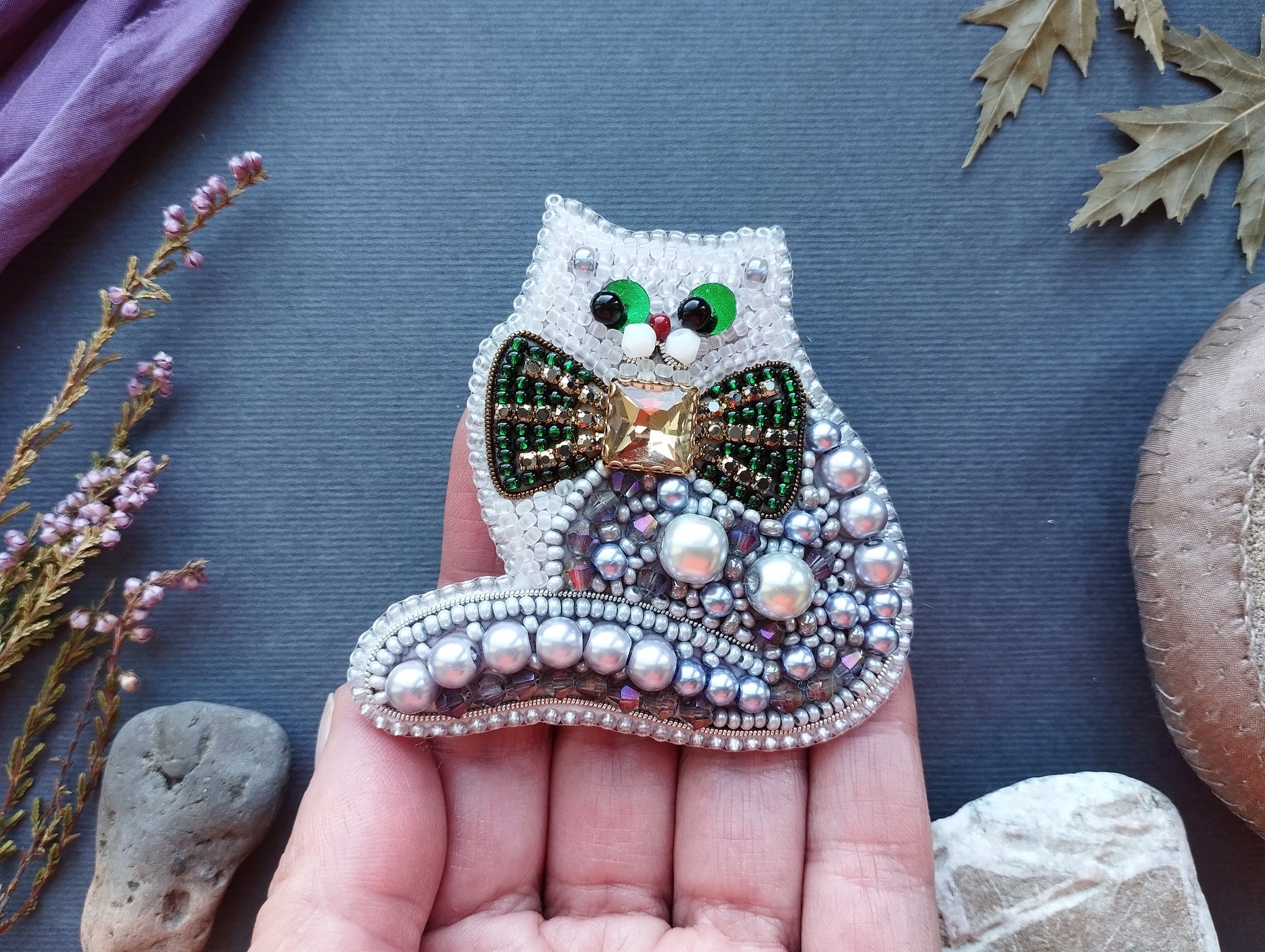 A hand holding a decorative beaded cat brooch with a bow tie, surrounded by various natural elements such as leaves, flowers, and rocks.