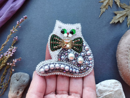 A hand holding a decorative beaded cat brooch with a bow tie, surrounded by various natural elements such as leaves, flowers, and rocks.