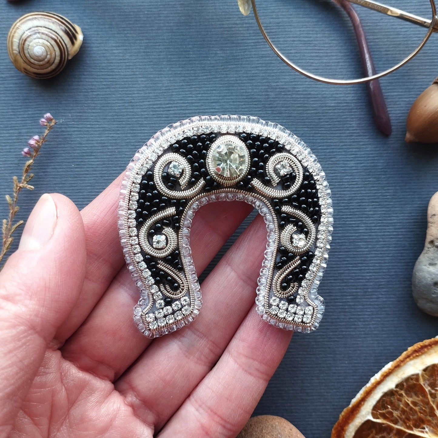A hand holding a decorative horseshoe-shaped brooch with a large gemstone in the center, surrounded by intricate beading and designs.