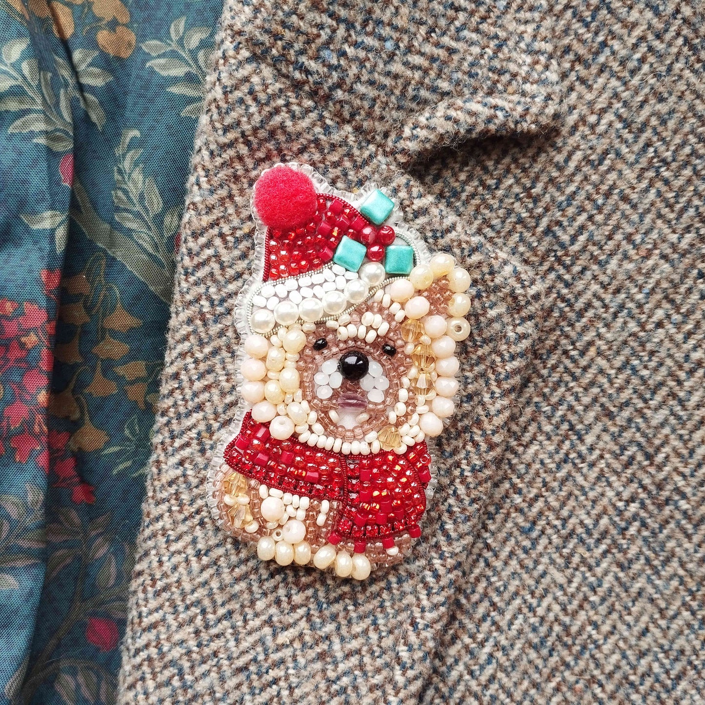 A close-up of a beaded, embroidered Christmas ornament in the shape of a dog wearing a red hat and scarf, placed on a tweed jacket.