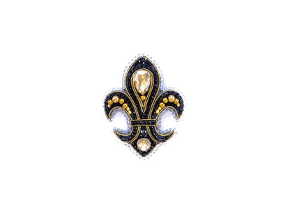 A decorative black and gold emblem featuring a fleur-de-lis design with a central gemstone surrounded by smaller gemstones and pearls.