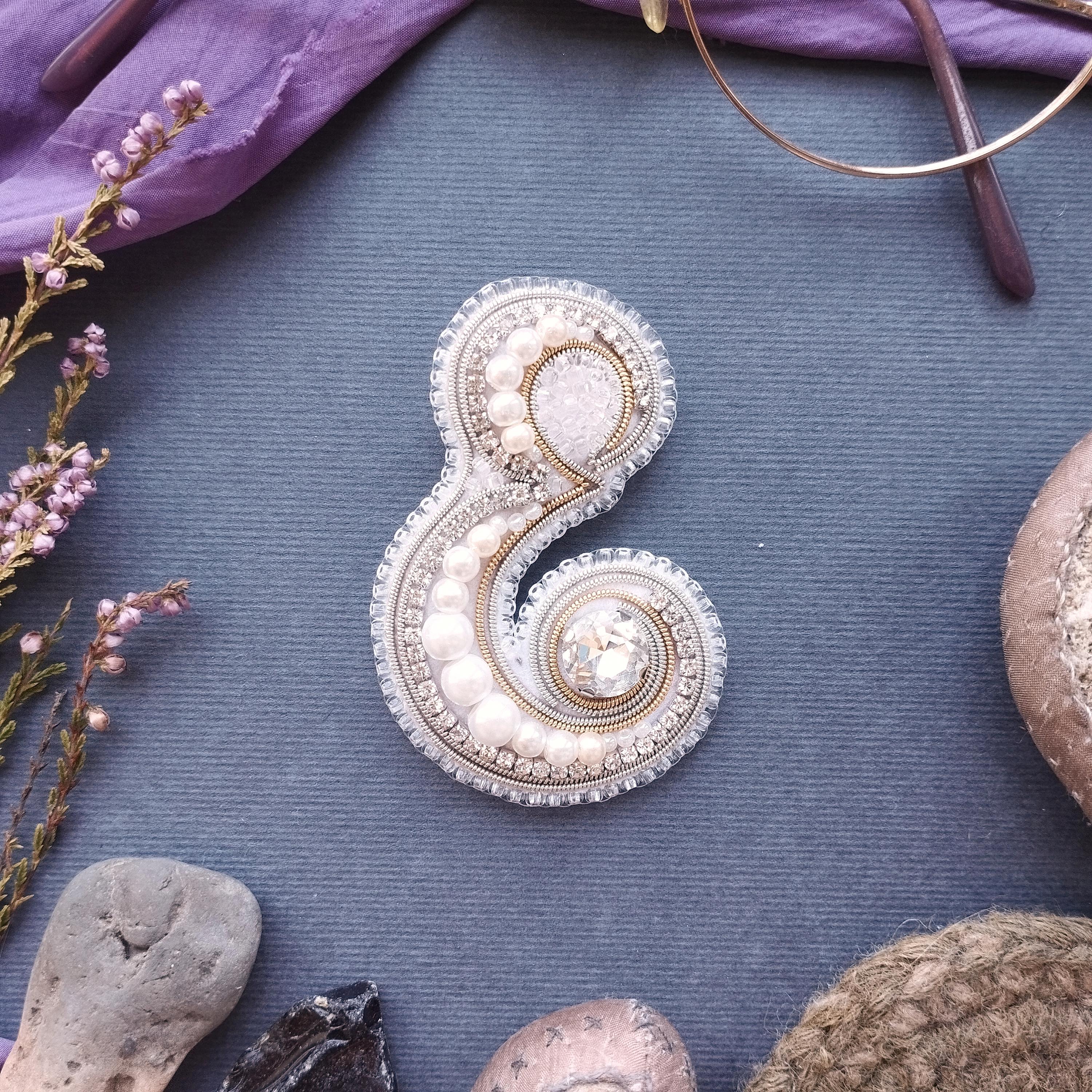 a decorative piece of jewelry, possibly a brooch or pendant, with intricate beading and pearl details, placed on a blue surface surrounded by various objects such as a pair of glasses, a plant, and a rock.