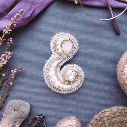 a decorative piece of jewelry, possibly a brooch or pendant, with intricate beading and pearl details, placed on a blue surface surrounded by various objects such as a pair of glasses, a plant, and a rock.