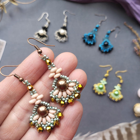 A hand holding a pair of beaded earrings with a floral design, surrounded by various other earrings and jewelry items on a gray background.