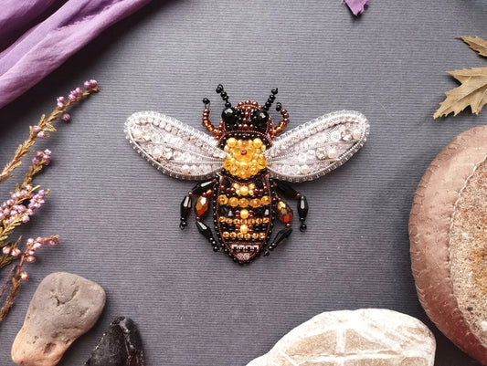 a detailed, hand-made beaded bee brooch with intricate designs and patterns, including a yellow and black color scheme. The brooch is placed on a gray background, surrounded by various natural elements such as leaves, rocks, and flowers.