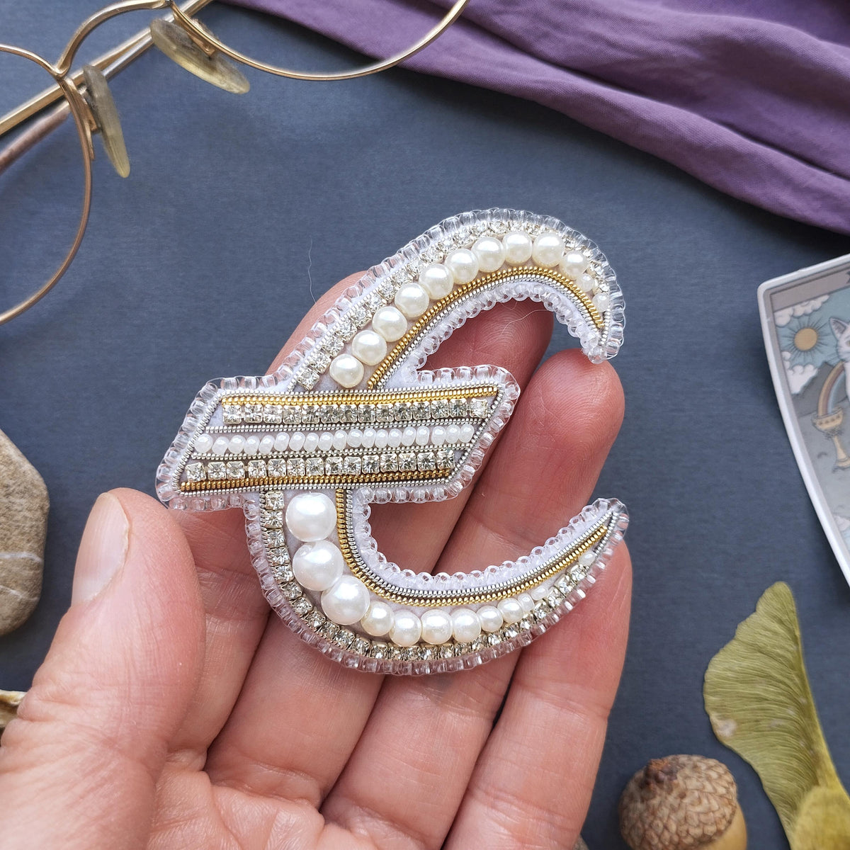 A hand holding a decorative piece of jewelry, which appears to be a brooch or pin, made of pearls and diamonds.