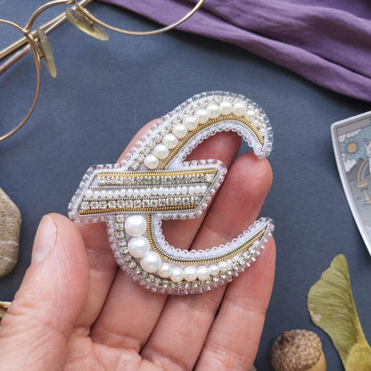 A hand holding a decorative piece of jewelry, which appears to be a brooch or pin, made of pearls and diamonds.